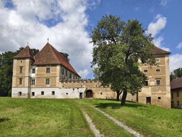 A historic castle with a red roof stands on a green meadow under a blue sky with clouds Styria Austria