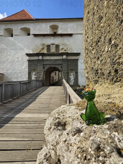 A glass frog sits on a stone in front of a fortress with a wooden bridge and blue sky Riegersburgs Styria Austria