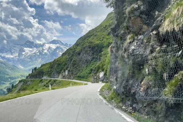 View of alpine road pass road south ramp from to Timmelsjoch Passo del Rombo with right at roadside of mountain road steep vertical rock is protected with strong wire chicken wire against rockfall, left terrain next to carriageway falls steeply into the depth, in the background partly snow-covered mountain peaks, South Tyrol, Italy