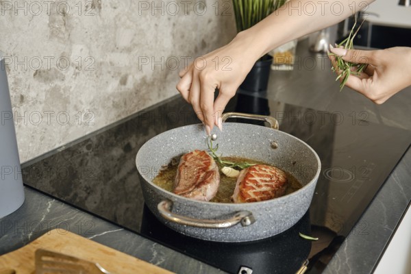 A person is preparing two pieces of duck breast in a frying pan, adding fresh herbs to enhance flavour. The kitchen features a contemporary design with a stone countertop and vibrant green plants