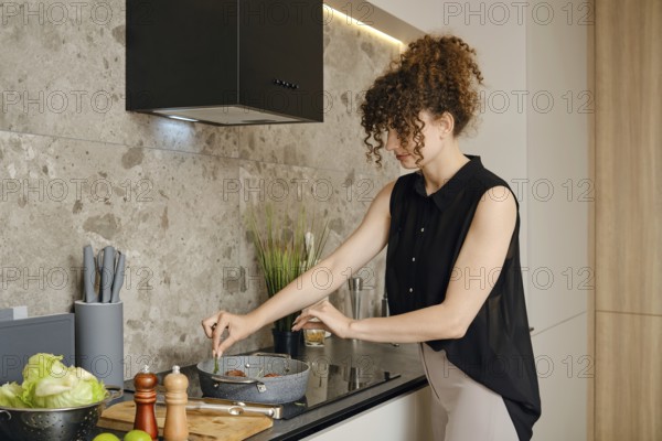 In a contemporary kitchen setup, a young woman focuses on preparing a meal. She adds fresh herbs in a frying pan with roasted meat