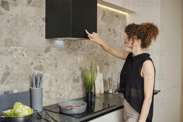 A woman prepares a healthy meal in a stylish kitchen, adjusting the vent fan above her cooking area. Fresh vegetables and a pot of sauce simmer on a sleek stovetop as she focuses on her task