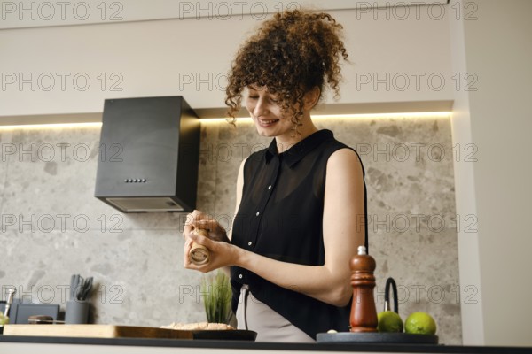 A woman stands in a stylish kitchen, smiling as she seasons a dish with a spice grinder. The contemporary design features sleek cabinets and a warm atmosphere, creating a lively cooking environment
