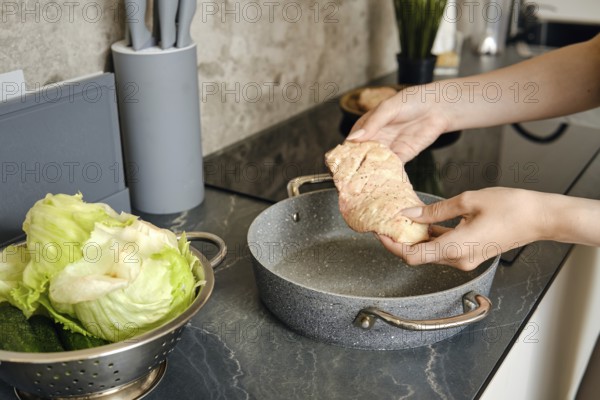 Hands gently hold a piece of duck above a cooking pot in a contemporary kitchen. Nearby, a bowl of fresh lettuce and cucumbers adds a vibrant touch, enhancing the fresh cooking atmosphere