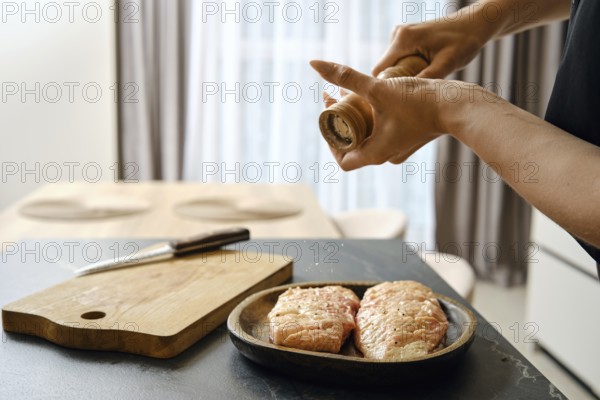 A person seasons fresh duck fillets with sea salt in a stylish kitchen. The wooden cutting board and knife are nearby, while natural light brightens the space from the window