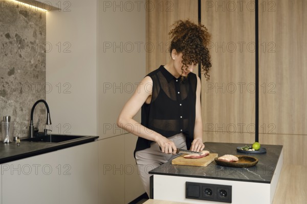 A woman with curly hair is cutting raw meat in a kitchen. Natural light illuminates her workspace as she focuses on meal preparation, showcasing a healthy cooking routine