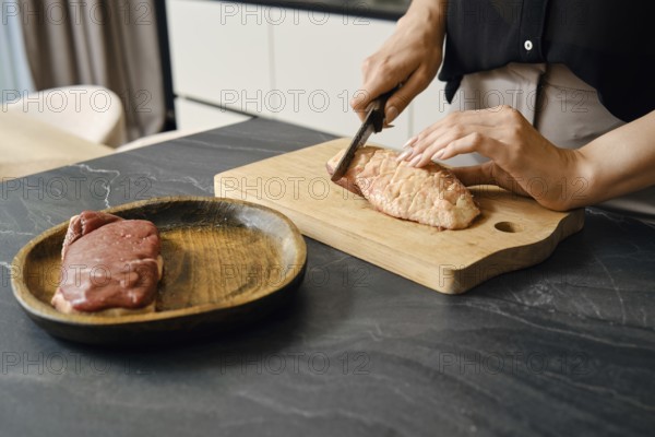 A person skillfully cuts the skin crosswise on a duck breast on a wooden board placed on a dark countertop. Another piece of meat rests on a wooden plate nearby