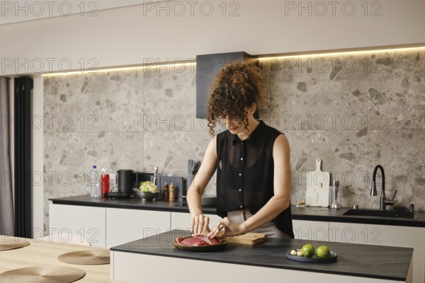 A woman takes duck meat from a wooden plate in a sleek modern kitchen. The kitchen features minimalist design elements with natural lighting and fresh ingredients nearby