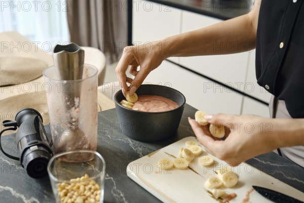A person adds slices of bananas to a creamy smoothie mixture in a black bowl. Nearby, a blender and a bowl of pine nuts suggest healthy meal prep