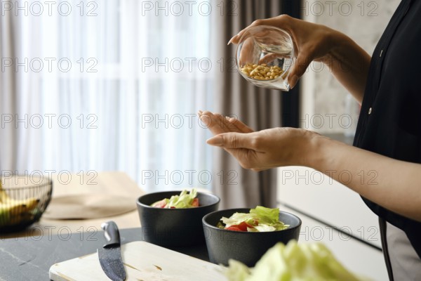A person adds pine nuts to a bowl filled with fresh salad ingredients, including lettuce and tomatoes, in a modern kitchen