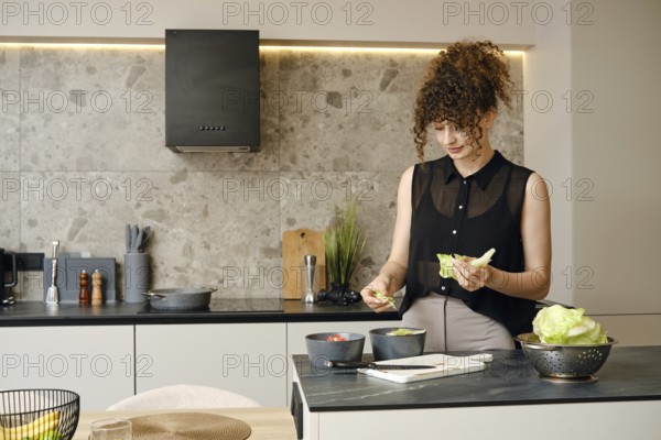 Woman with curly hair prepares a fresh salad in a contemporary kitchen. She is tearing lettuce with her hands and mixing ingredients. The kitchen features stylish decor and modern appliances