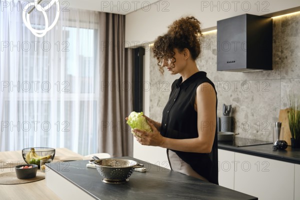 In a contemporary kitchen filled with natural light, a woman holds a head of lettuce while preparing to create a fresh salad. Various fruits and vegetables are seen nearby