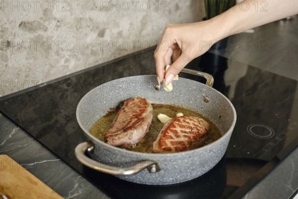A hand adds garlic to sizzling duck steaks in a pan on the induction stovetop. The preparation showcases a focus on culinary skills and the art of cooking meat perfectly