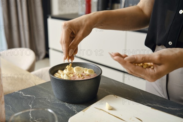 In a modern kitchen, a person adds pine nuts over sliced bananas to a bowl filled with a smoothie base. The countertop has fresh ingredients, highlighting a healthy breakfast preparation