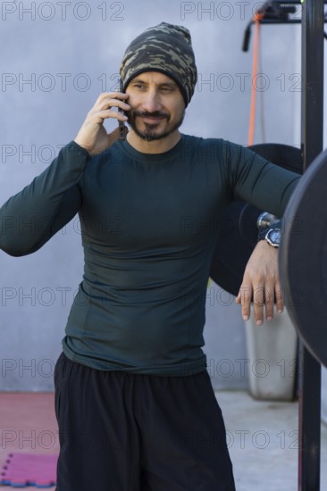 Athlete takes a break from his workout, leaning against a weightlifting rack and talking on his cell phone, demonstrating a blend of fitness and connectivity