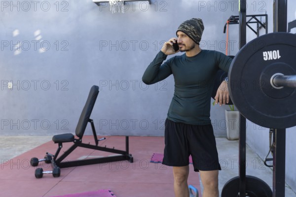 Athlete taking a break from an intense workout, leaning against a weightlifting rack while engaged in a conversation on his mobile phone in a home gym setting