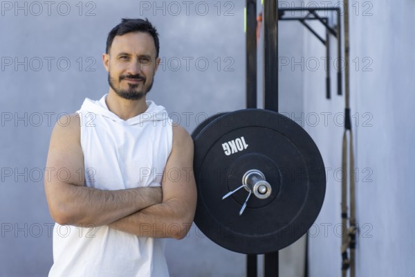 Muscular personal trainer with crossed arms leans confidently against a weight plate in an outdoor gym, radiating expertise and dedication to fitness and strength training