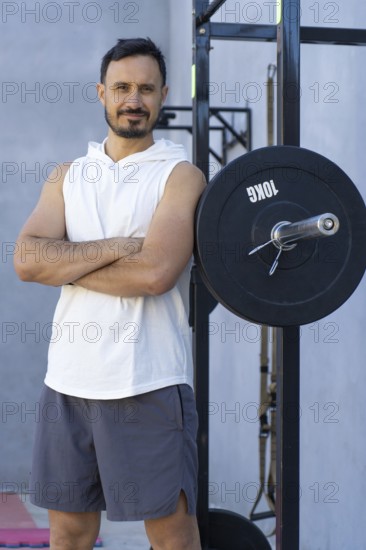 Sporting a confident expression, a sportsman with crossed arms leans against a weight plate in an outdoor gym, embodying strength and determination in his fitness journey