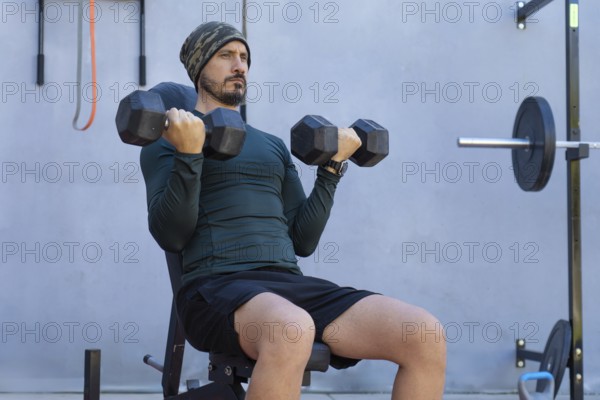 Focused male athlete sitting on a weight bench, lifting dumbbells and performing bicep curls during an intense outdoor workout routine, showcasing dedication and commitment to fitness