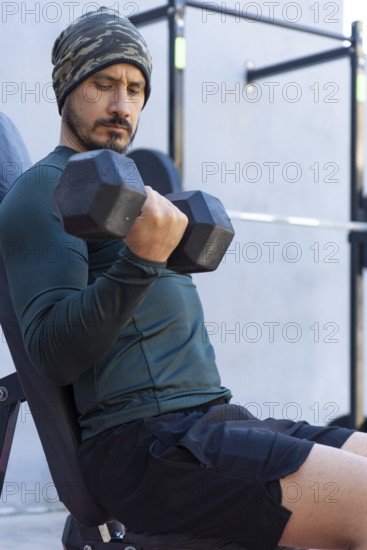 Focused athlete lifts dumbbells, performing bicep curls while seated on a bench in an outdoor gym, demonstrating strength and dedication to fitness