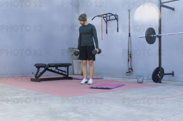 Young sportsman lifting two black dumbbells while engaging in a workout in a private outdoor gym, showcasing dedication to fitness and strength training in a personal space