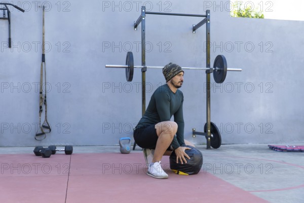 Young sportsman wearing beanie and thermal shirt performing wall ball squats in an outdoor gym, demonstrating strength and fitness with gym equipment in the background