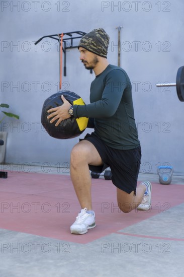 Athlete wearing a beanie, long-sleeved shirt, and shorts performs lunges while holding a medicine ball in an outdoor gym setting, demonstrating strength and balance