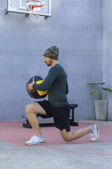 Young sportsman wearing a beanie and sportswear is lunging with a medicine ball in an outdoor gym, positioned near a basketball hoop in an urban setting