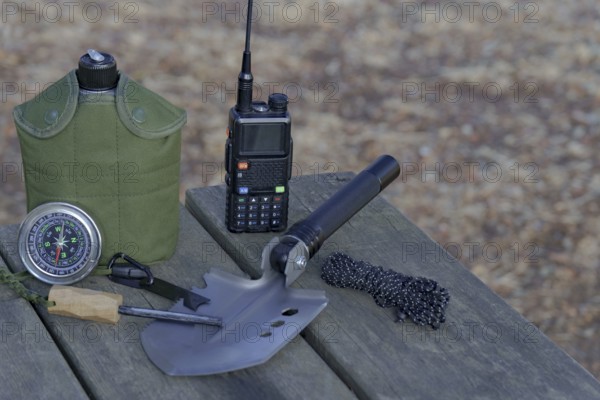 Essential survival gear including walkie talkie, compass, folding shovel, and fire starter are arranged on a wooden table in nature