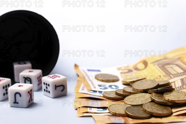 White dice with black letters and red dots are falling from a black dice cup, lying near some 50 euro banknotes and coins