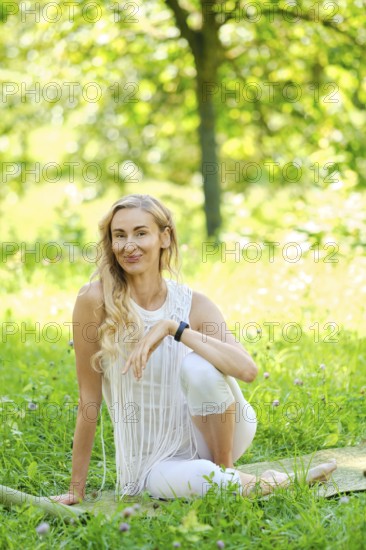 A woman dressed in white sits gracefully on a mat in a lush park, surrounded by vibrant green grass and blooming flowers. She enjoys a peaceful moment during her yoga practice under the warm sun