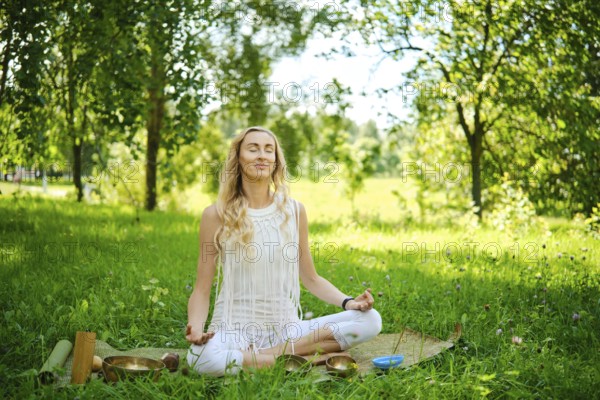 A woman sits cross-legged on a mat in a lush green park, eyes closed and arms resting on her knees. She focuses on mindfulness and tranquility, surrounded by trees and nature