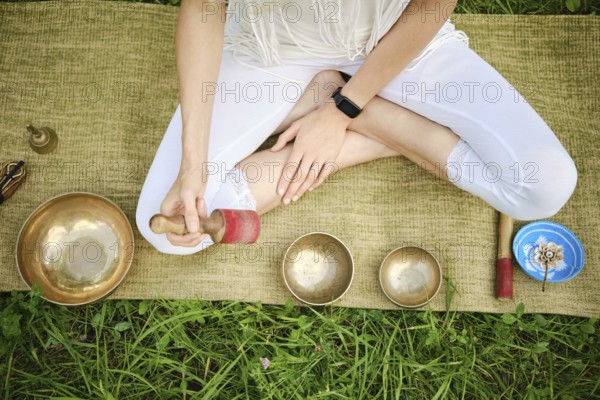 Overhead view of an unrecognizable person sitting cross-legged on a mat in a serene outdoor environment, gently playing singing bowls