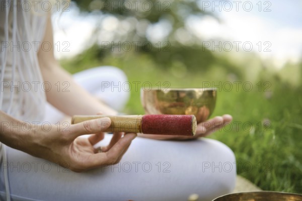 A person is holding a red-tipped mallet next to a singing bowl, seated on grass in a serene outdoor setting. This meditative activity takes place in a tranquil environment, encouraging relaxation