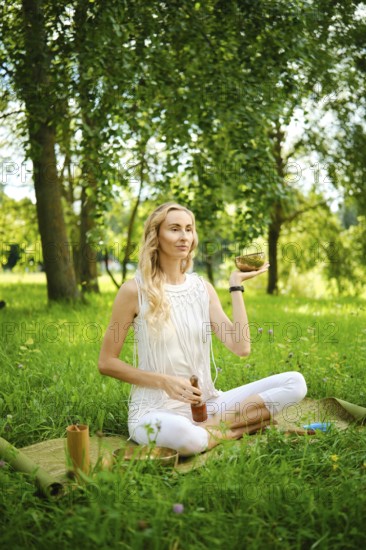 A woman practices meditation outdoors, holding a singing bowl while seated on a mat in a field. The surrounding nature and sunlight enhance the calming atmosphere of the moment