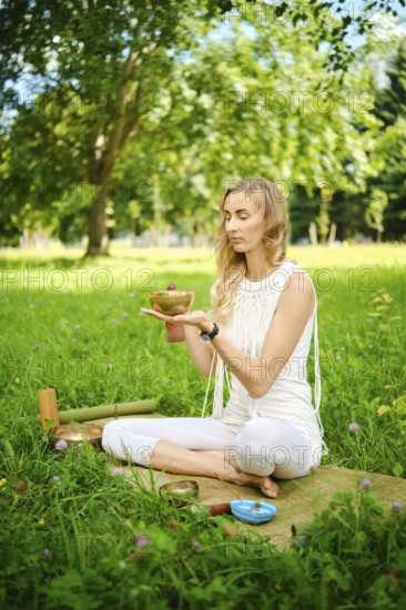 A woman sits cross-legged in a grassy park, holding a singing bowl while meditating. Surrounded by lush trees and a serene environment, she focuses on sound healing and inner peace