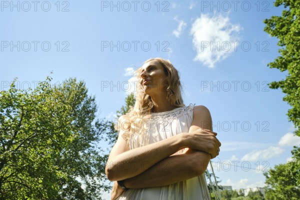 In a green park on a bright day, a woman stands with her arms crossed, gazing upwards. Sunlight highlights her long hair and flowing dress, creating a serene atmosphere