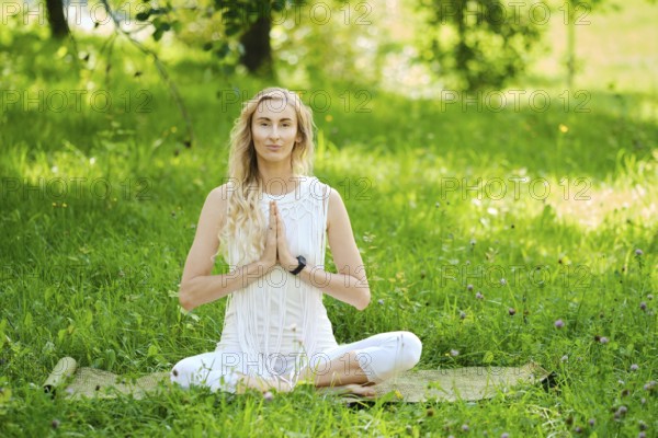 A woman dressed in white sits cross-legged on a mat, practicing yoga outdoors. Surrounded by vibrant green grass and trees, she has her hands in a prayer position, embodying peace and tranquility