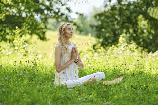 A woman sits cross-legged on soft grass in a vibrant green field. She closes her eyes and brings her hands together in a prayer position, embracing the tranquility of nature on a sunny day