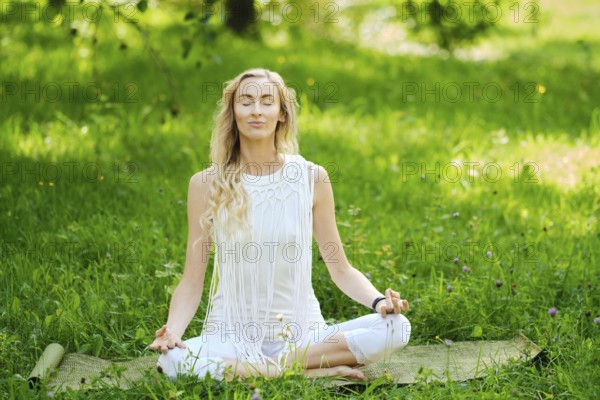 A woman in a white outfit sits cross-legged on a mat, eyes closed and hands resting on her knees in a peaceful meditation. Lush grass and trees surround her, creating a tranquil atmosphere