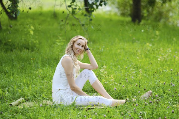A young woman sits comfortably on a blanket in a lush green park. She has wavy hair and wears a white outfit