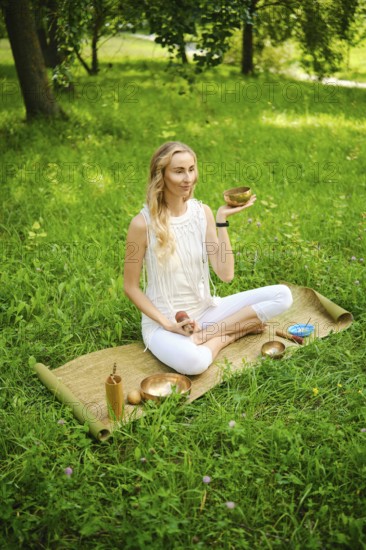 A woman engages in a calming meditation session, holding a singing bowl while seated on a mat in a lush green environment. The peaceful atmosphere enhances her focus on mindfulness