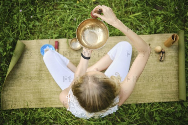 Overhead view of a woman sitting cross-legged on a mat, skillfully playing a singing bowl. Surrounded by lush grass, the tranquil atmosphere enhances the meditative experience