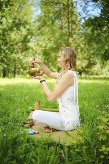 A woman sits cross-legged on a mat in a lush green field, playing a singing bowl with a mallet. Sunlight filters through the trees as she engages in meditation and mindfulness