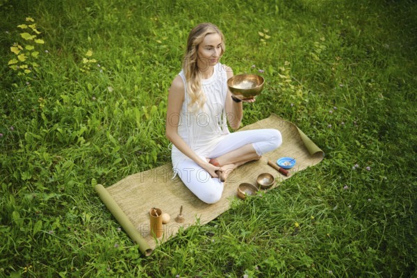 A woman sits cross-legged on a mat in a lush green field, holding a singing bowl. She is fully engaged in a meditative practice, surrounded by additional bowls and calming symbols