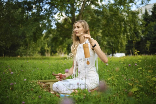 A woman sits cross-legged on the grass, surrounded by greenery, gently striking singing bowls with mallets