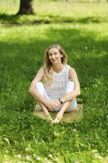 A barefoot woman sits on a mat in a vibrant green park, enjoying the sunny weather. She has long blonde hair and wears a light outfit, radiating tranquility and calmness in nature