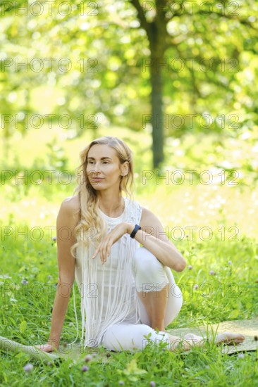 A woman with long hair sits elegantly in a vibrant green park. Dressed in white, she poses thoughtfully amidst flowers and sunshine while enjoying the peaceful atmosphere