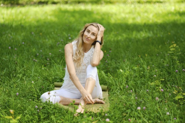 A young woman dressed in white relaxes on a mat in a lush green field. She smiles gently while resting her chin on her hand, surrounded by vibrant grass and wildflowers under a clear blue sky
