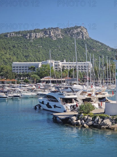 Kemer marina filled with numerous boats and yachts with Calis Hill in the background. A lighthouse stands prominently on the rocky pier, overlooking the clear blue water under a bright blue sky